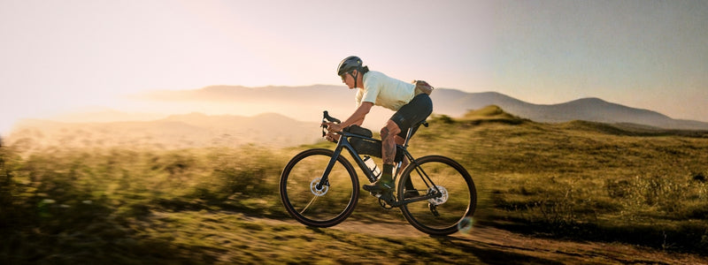 Kuvassa nainen pyöräilee gravel pyörällä maastossa, jossa takana on niittyä  ja vuoria