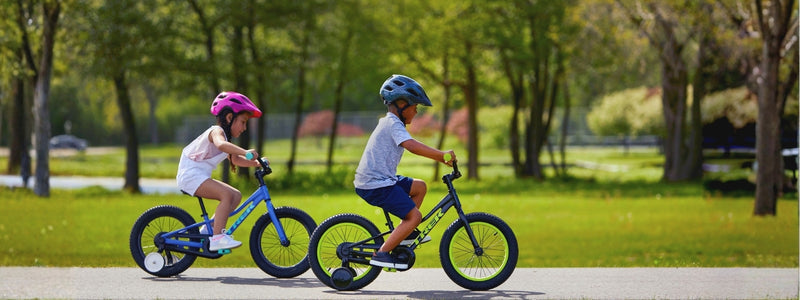 Kaksi pientä lasta pyöräilee apupyöräisillä pyörillä puiston pyörätiellä