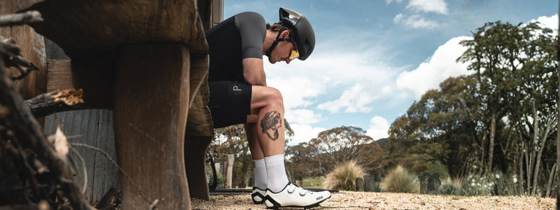 Miehellä on pyöräilyvarusteet päällä ja hän istuu penkillä, ja katsoo maahan