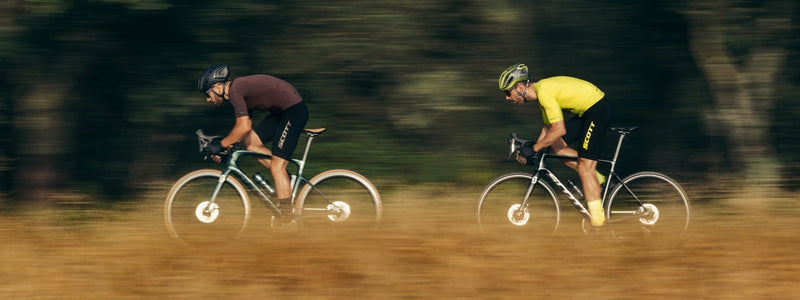 Kaksi miestä polkee kovaa vauhtia maantiellä, pellon vieressä